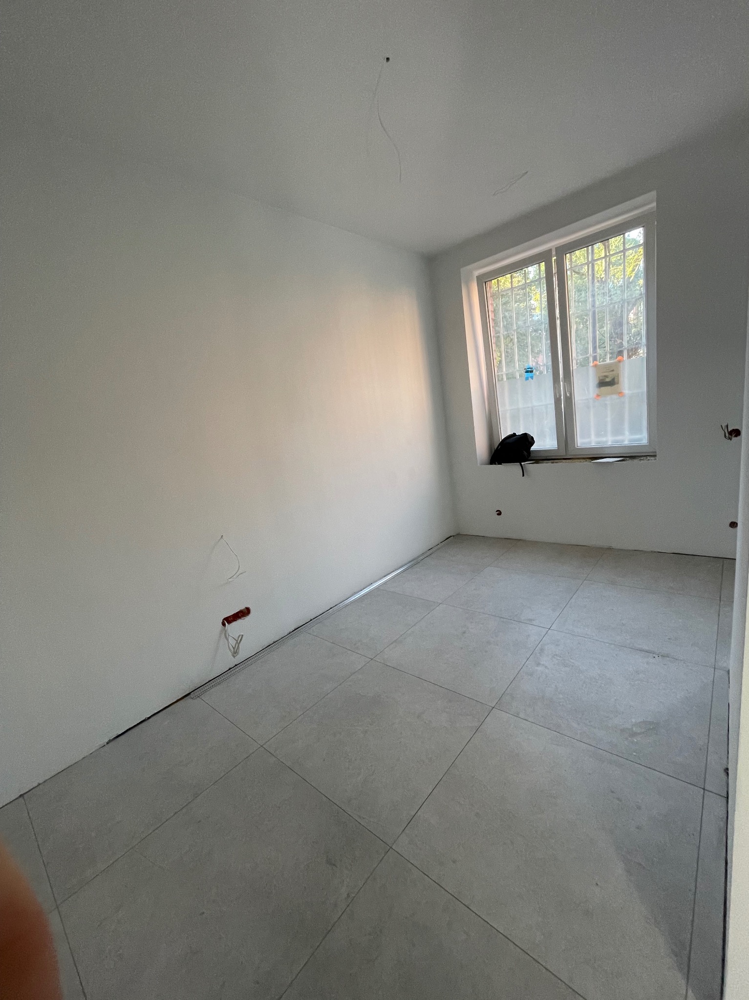 Wnętrze pokoju po remoncie z ułożoną szarą podłogą z płytek, świeżo pomalowanymi ścianami na biało i oknem z widokiem na zieleń.