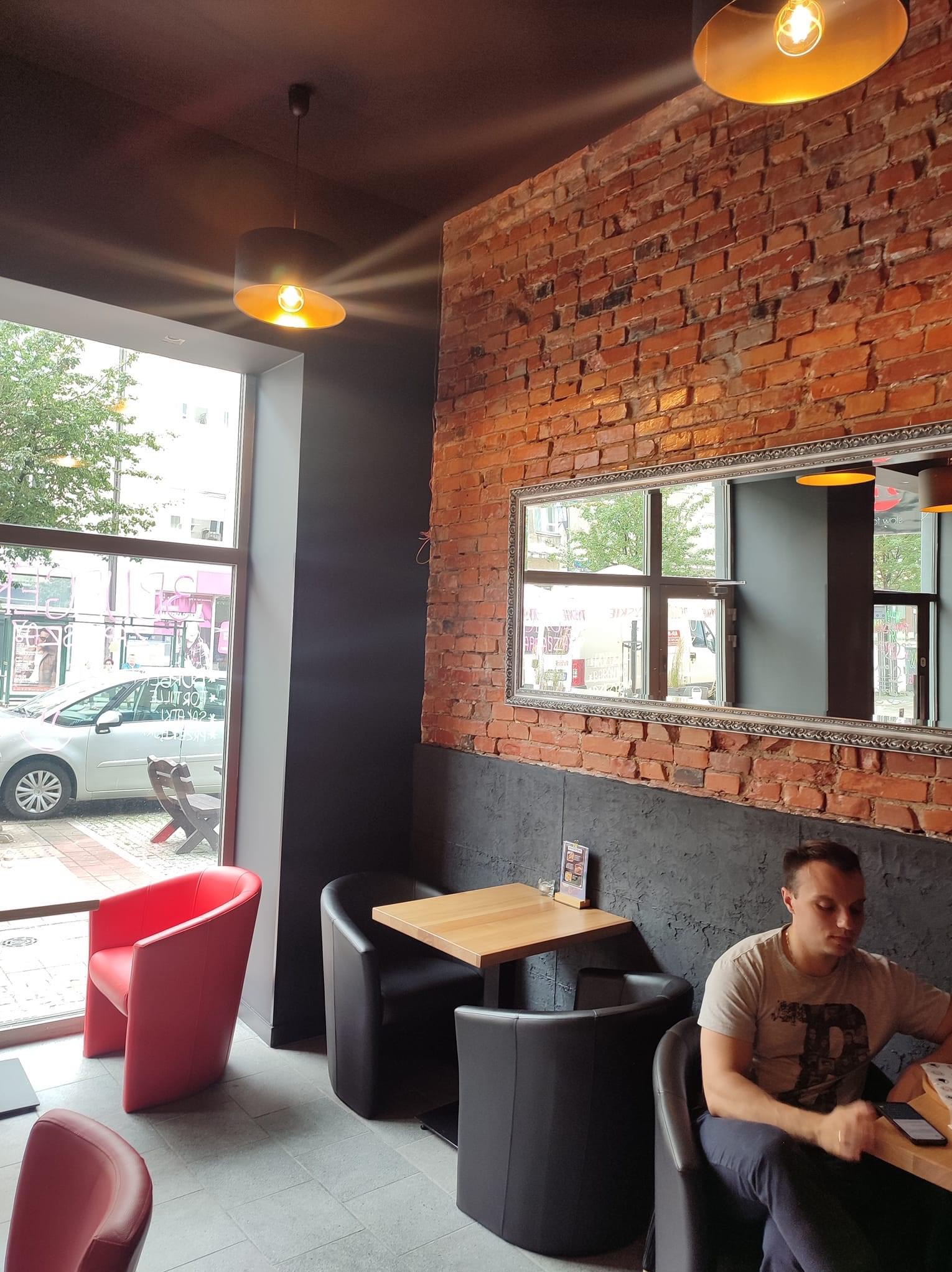 Wnętrze kawiarni z ceglaną ścianą, lustrem w ozdobnej ramie, drewnianymi stolikami i krzesłami w kolorze czarnym i czerwonym, oświetlone ciepłym światłem lamp wiszących.