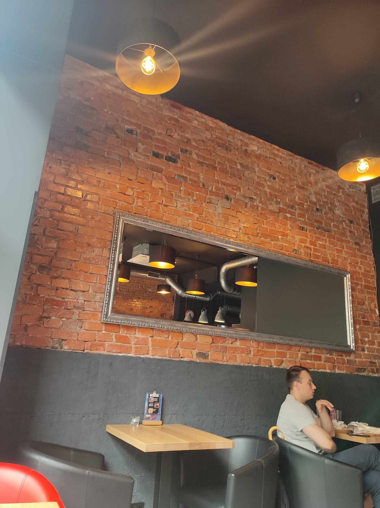 Wnętrze kawiarni z czerwoną cegłą na ścianie, dekoracyjnym lustrem w srebrnej ramie, widoczną instalacją sufitową i oświetleniem, oraz siedzącym mężczyzną przy stoliku.