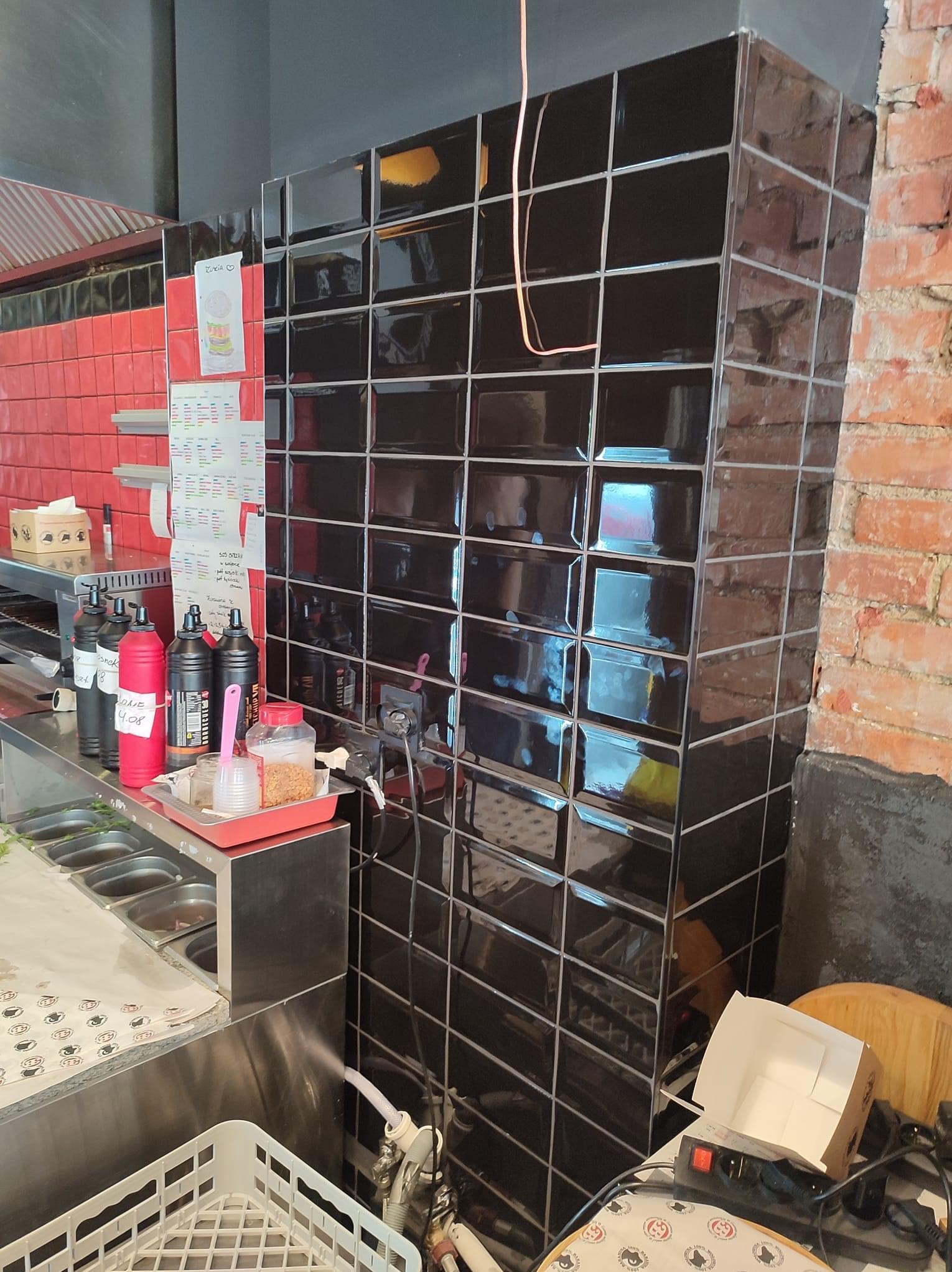 Ściana wyłożona czarnymi, błyszczącymi płytkami typu metro, przylegająca do ściany z czerwonej cegły w lokalu gastronomicznym, widoczne elementy wyposażenia kuchni i menu na ścianie.