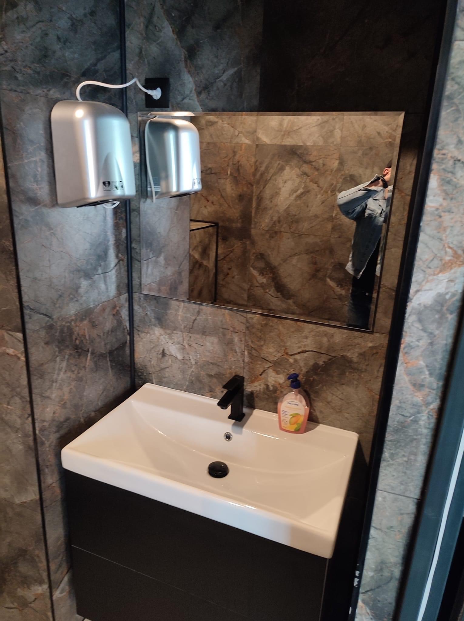 Nowoczesna łazienka z umywalką nablatową, czarną armaturą, marmurowymi płytkami na ścianach, dwoma srebrnymi suszarkami do rąk i odbiciem osoby w lustrze.