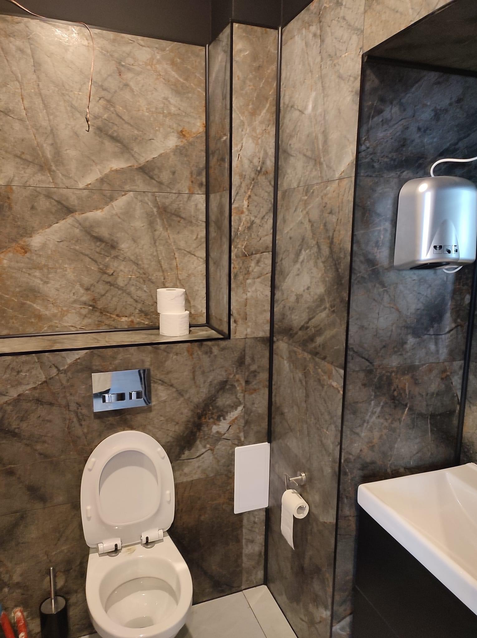 Nowoczesna łazienka z marmurowymi płytkami, wiszącą toaletą z otwartą klapą, srebrnym podajnikiem na ręczniki papierowe i rolką papieru toaletowego na ścianie.