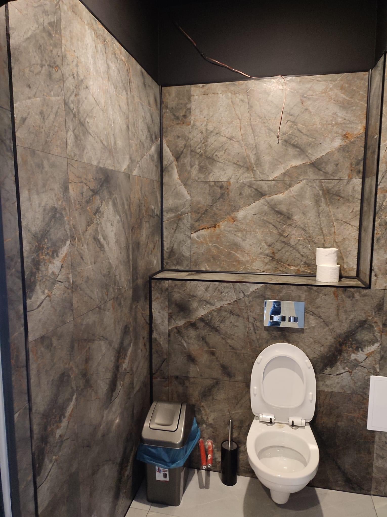 Nowoczesna toaleta w łazience wykończonej płytkami imitującymi marmur, z widocznym fragmentem niedokończonej instalacji elektrycznej pod sufitem.