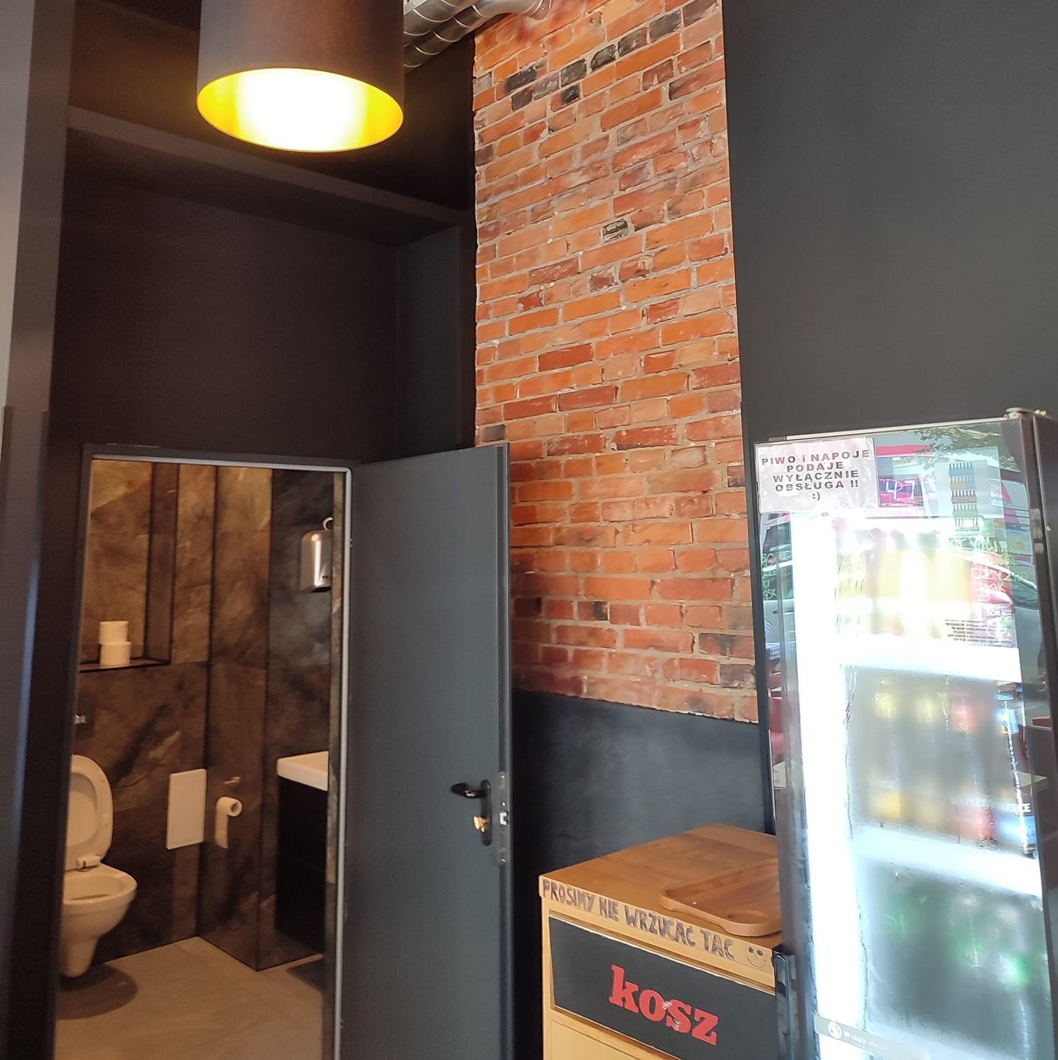 Wnętrze lokalu gastronomicznego z widokiem na toaletę z szarą, otwartą ościeżnicą, ścianą z czerwonej cegły i lodówką z napojami.