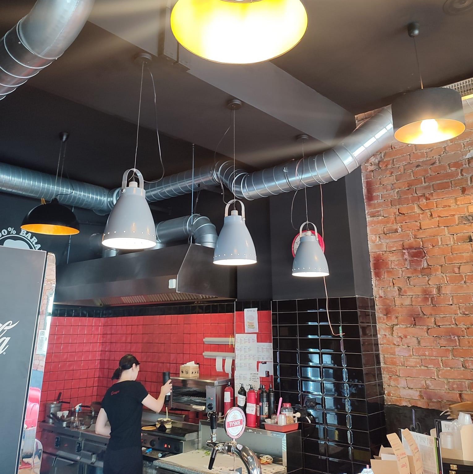 Wnętrze lokalu gastronomicznego w trakcie pracy: widoczna kuchnia z czerwonymi płytkami, czarne płytki na filarze, ceglana ściana, lampy wiszące i system wentylacji pod sufitem.