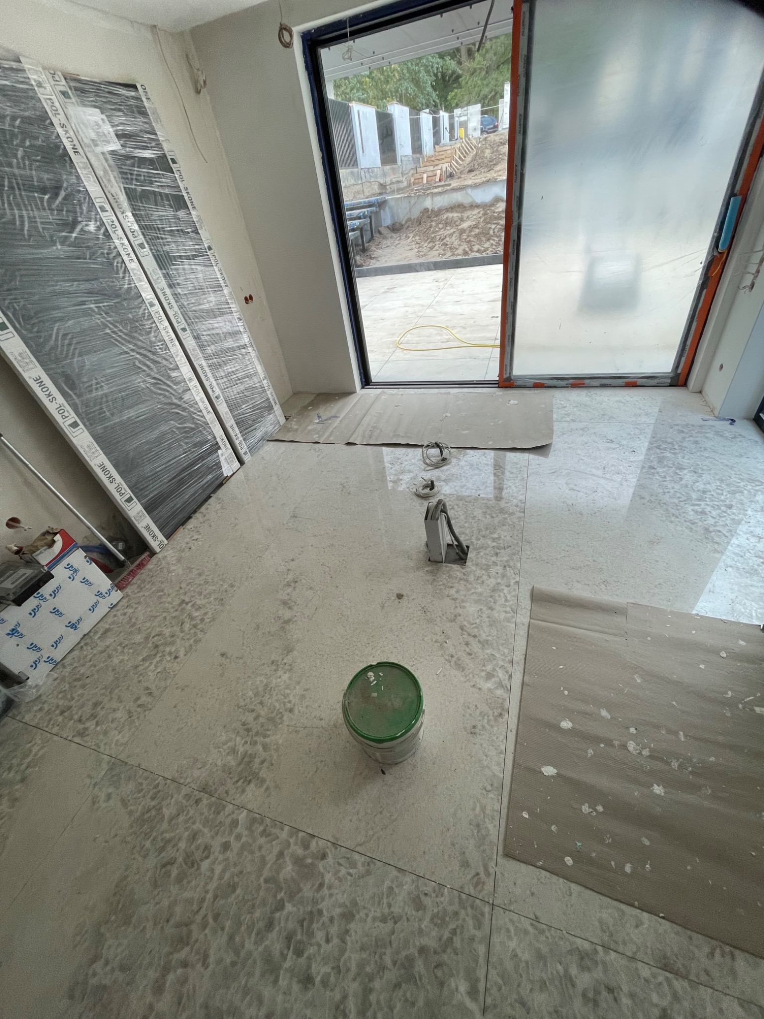 Wnętrze w trakcie prac wykończeniowych: położona jasna, marmurkowa glazura podłogowa, widoczne drzwi w folii ochronnej oparte o ścianę, puszka z farbą i narzędzia glazurnicze.