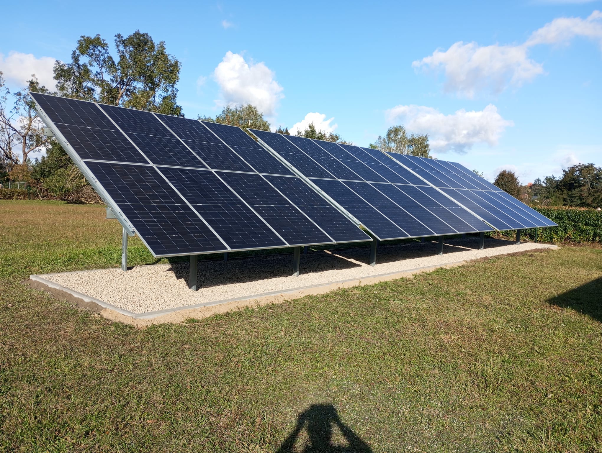 Rząd paneli słonecznych ustawionych na trawniku z podsypką z jasnego żwiru i widocznym cieniem fotografa w dolnej części kadru.