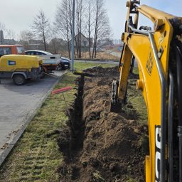 Usługi Koparko-Ładowarką, Minikoparką, Transport - Żółta koparka JCB częściowo w kadrze, wykopany wąski rów w ziemi, obok trawa i chodnik. W tle zaparkowane pojazdy i budynki. Widok w słoneczny dzień.