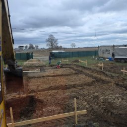 Usługi Koparko-Ładowarką, Minikoparką, Transport - Żółta koparka przy wykopie pod fundamenty z widocznym zbrojeniem, pracownikami i kontenerem w tle. Prace ziemne w toku, pochmurne niebo.