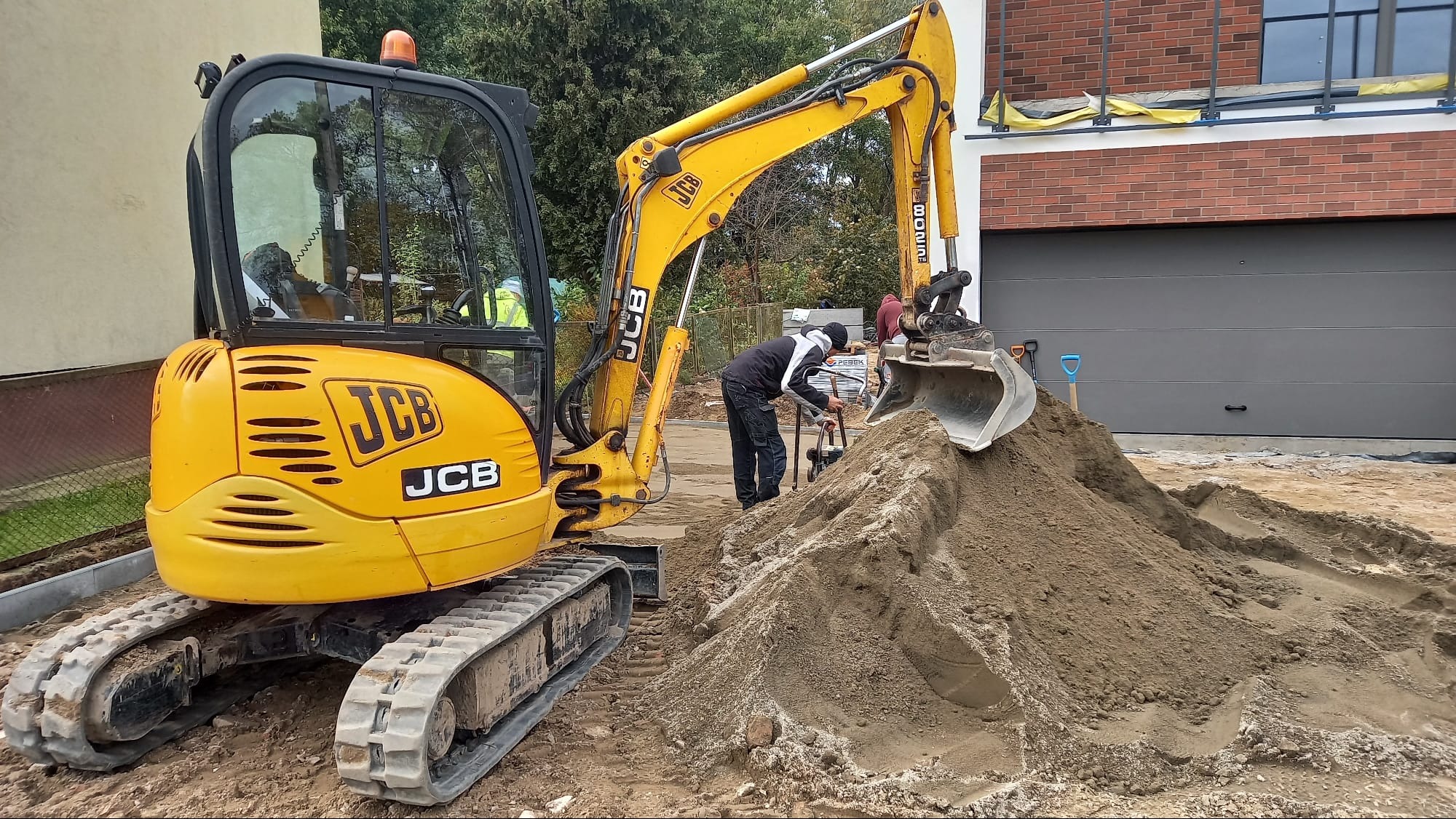 Żółta mini koparka JCB obok pryzmy piasku, w tle pracownik z ubijarką, nowy budynek z szarą bramą garażową oraz ułożona kostka brukowa.