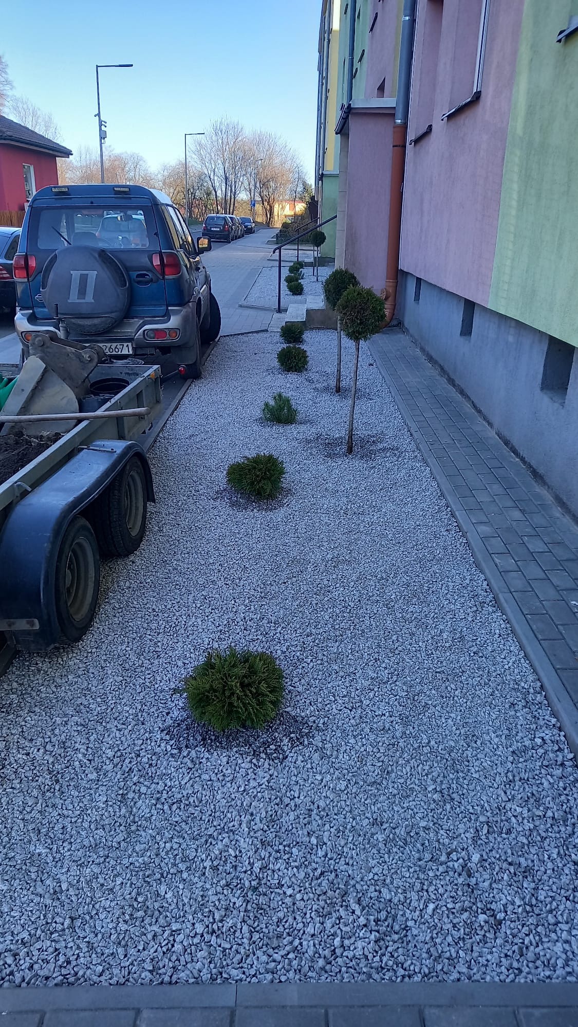 Świeżo wysypany żwir ozdobny z nasadzeniami młodych krzewów iglastych wzdłuż budynku mieszkalnego, przyczepa z resztkami ziemi i samochód terenowy w tle.