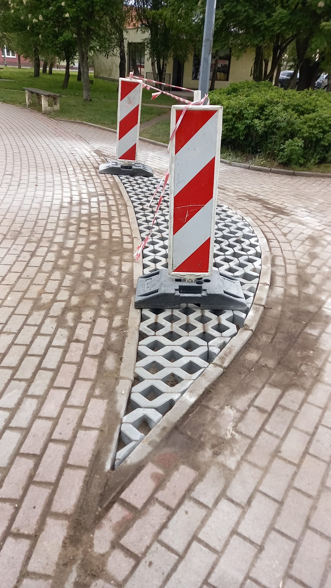 Fragment nowej nawierzchni z ażurowych płyt betonowych, oddzielonej od starej kostki barierkami z taśmą. Widoczne ślady ziemi i piasku.