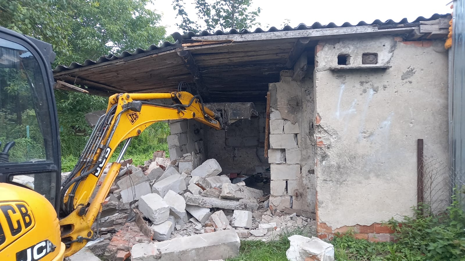 Żółta koparka JCB podczas częściowej rozbiórki murowanego budynku gospodarczego z zadaszeniem. Gruz leży przed budynkiem. Widoczne ślady uszkodzeń.