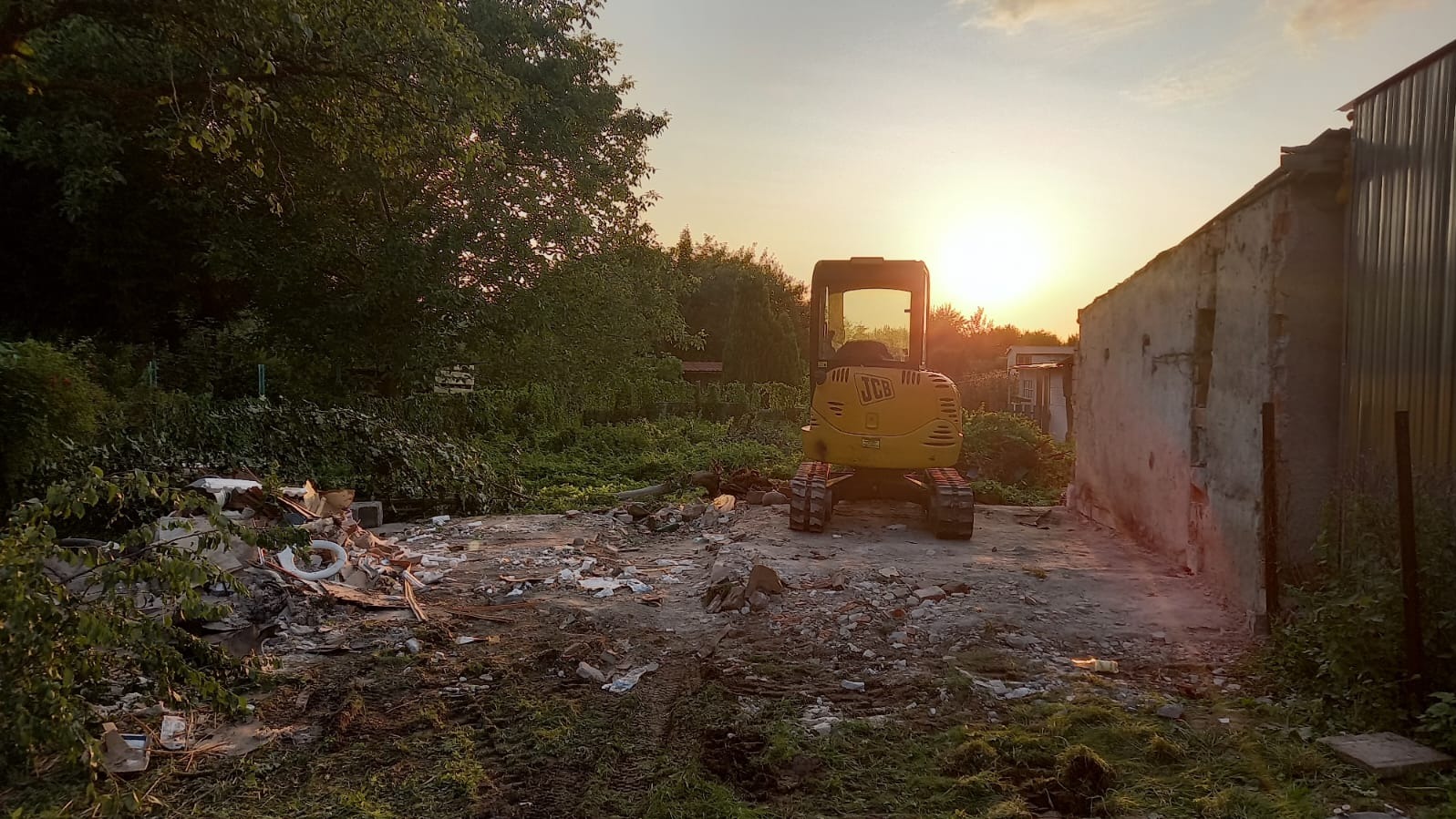 Żółta mini koparka JCB na terenie po rozbiórce, gruz i resztki materiałów budowlanych, zachód słońca w tle.
