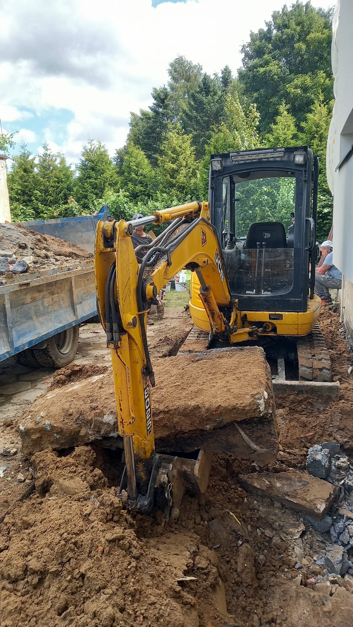 Żółta mini koparka JCB w trakcie prac ziemnych, usuwa fragmenty betonu. W tle widoczna przyczepa z gruzem, drzewa i fragment budynku.