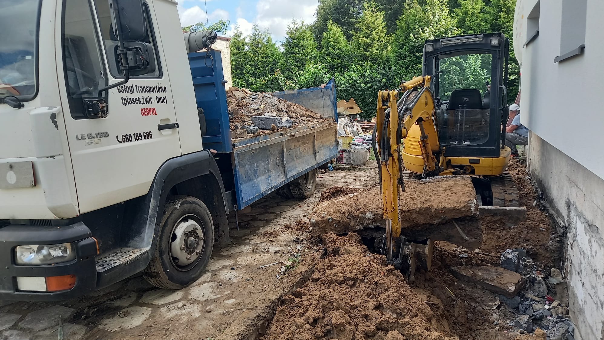 Żółta mini koparka i ciężarówka z gruzem podczas wykopu pod fundamenty w Sławnie. Widoczny operator koparki. Prace ziemne przy budynku.