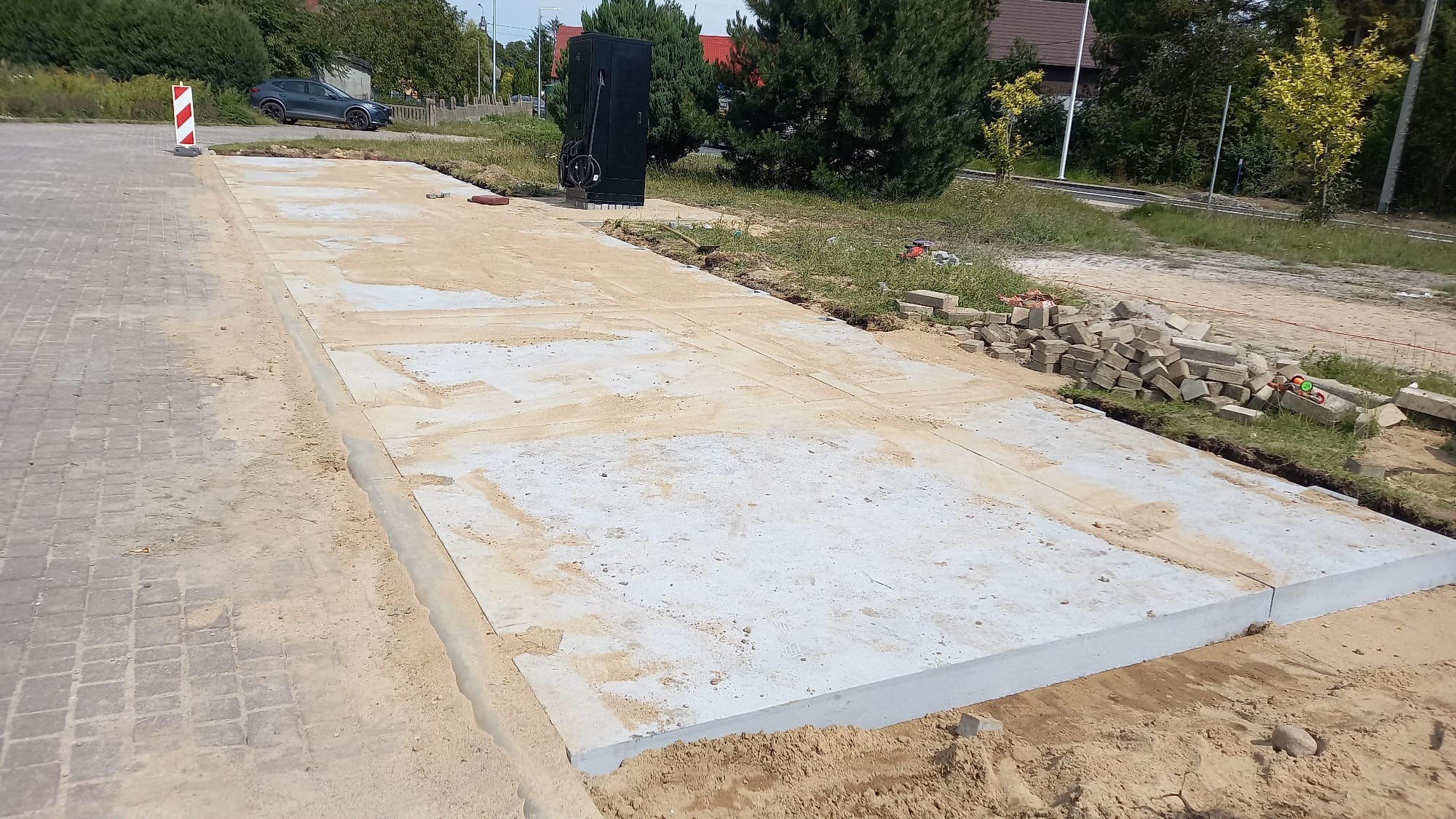 Przygotowany teren pod kostkę brukową z widocznymi obrzeżami i stertą kostki. Widoczne elementy infrastruktury i zieleń w tle. Ujęcie z góry.