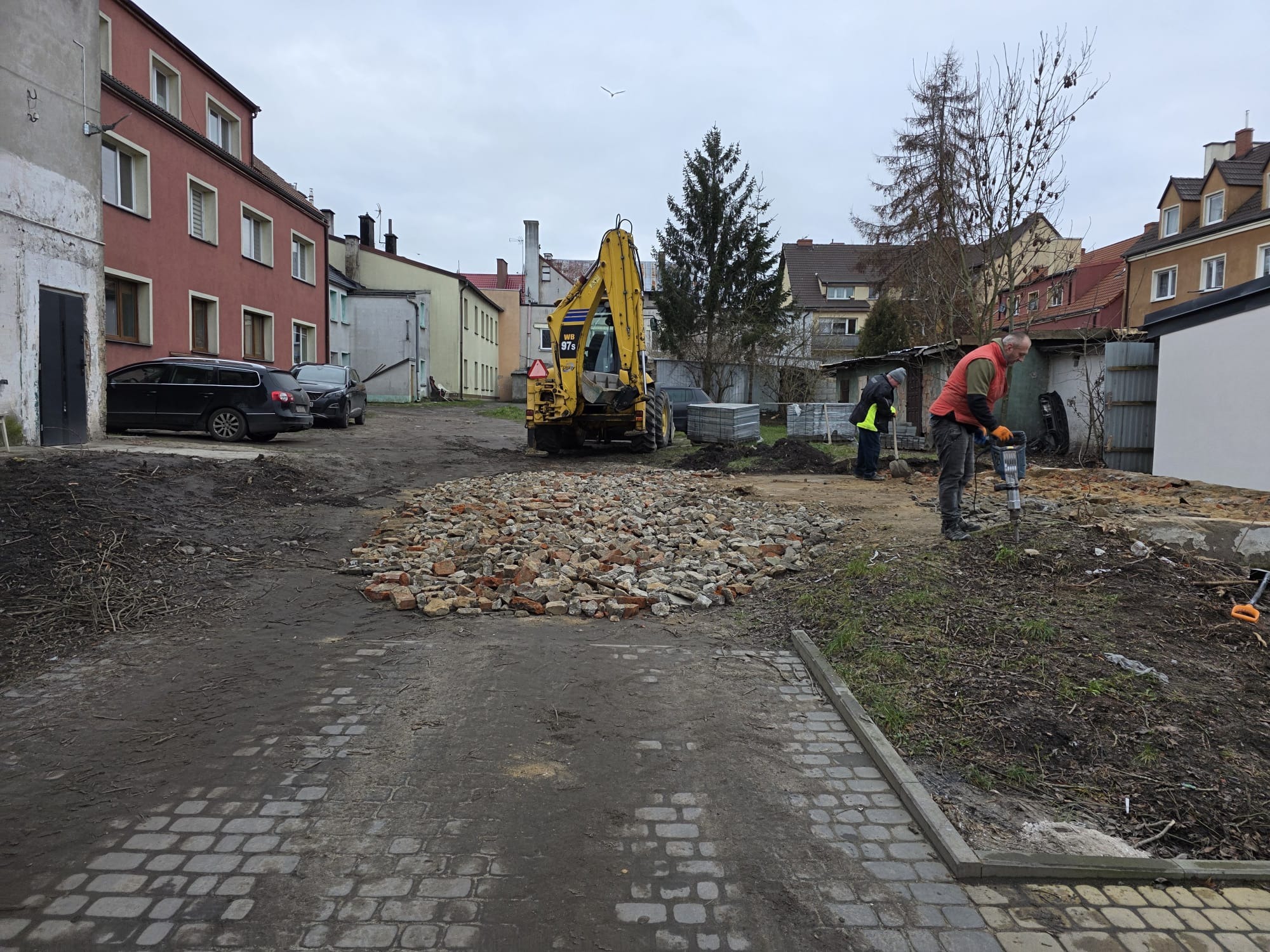 Demontaż starej kostki brukowej na posesji z użyciem młota pneumatycznego, w tle koparka i budynki mieszkalne w pochmurny dzień.