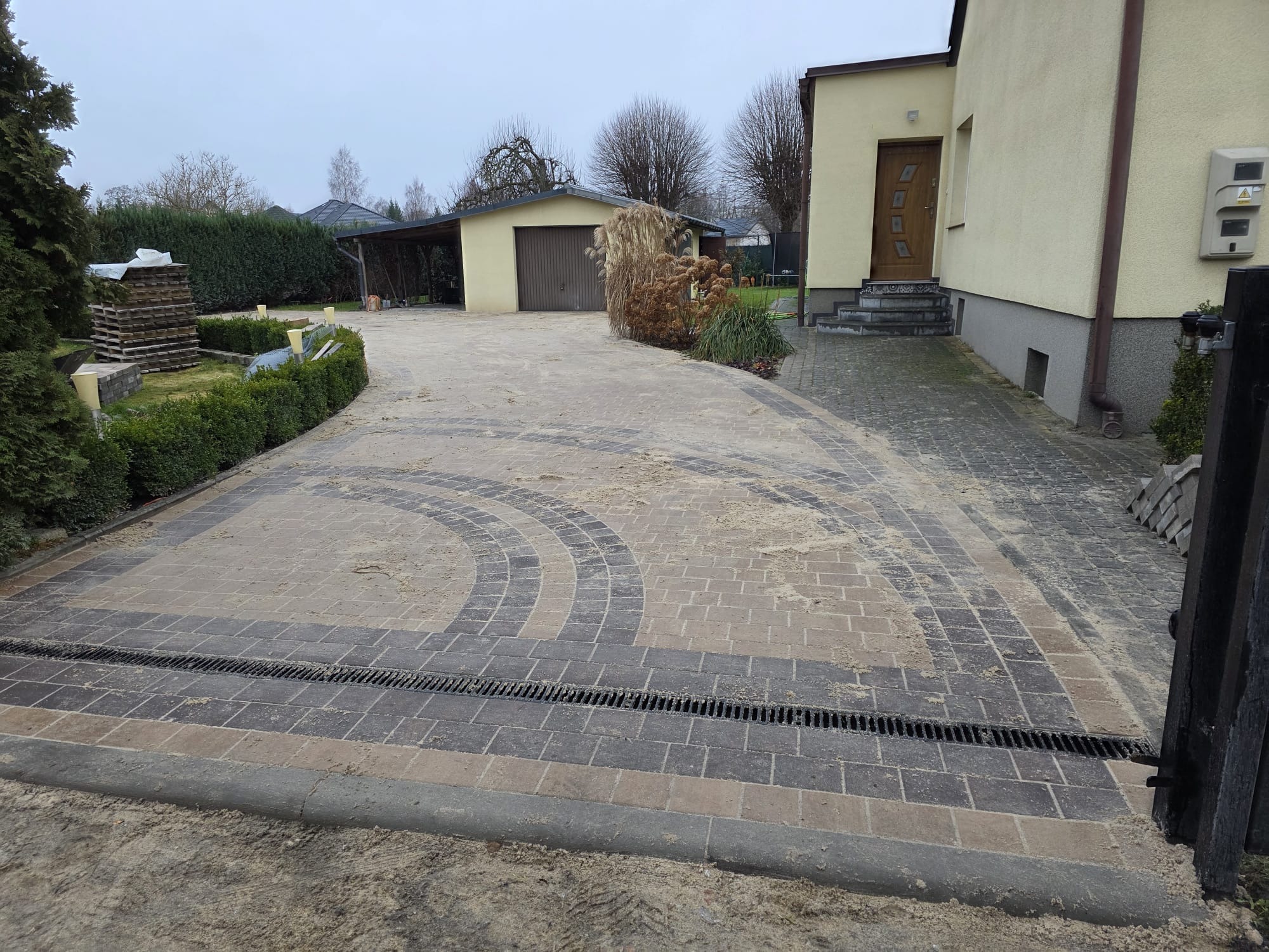 Ułożona kostka brukowa w odcieniach szarości i beżu tworząca wzór na podjeździe. Widoczny odpływ liniowy i fragment domu z garażem w tle.