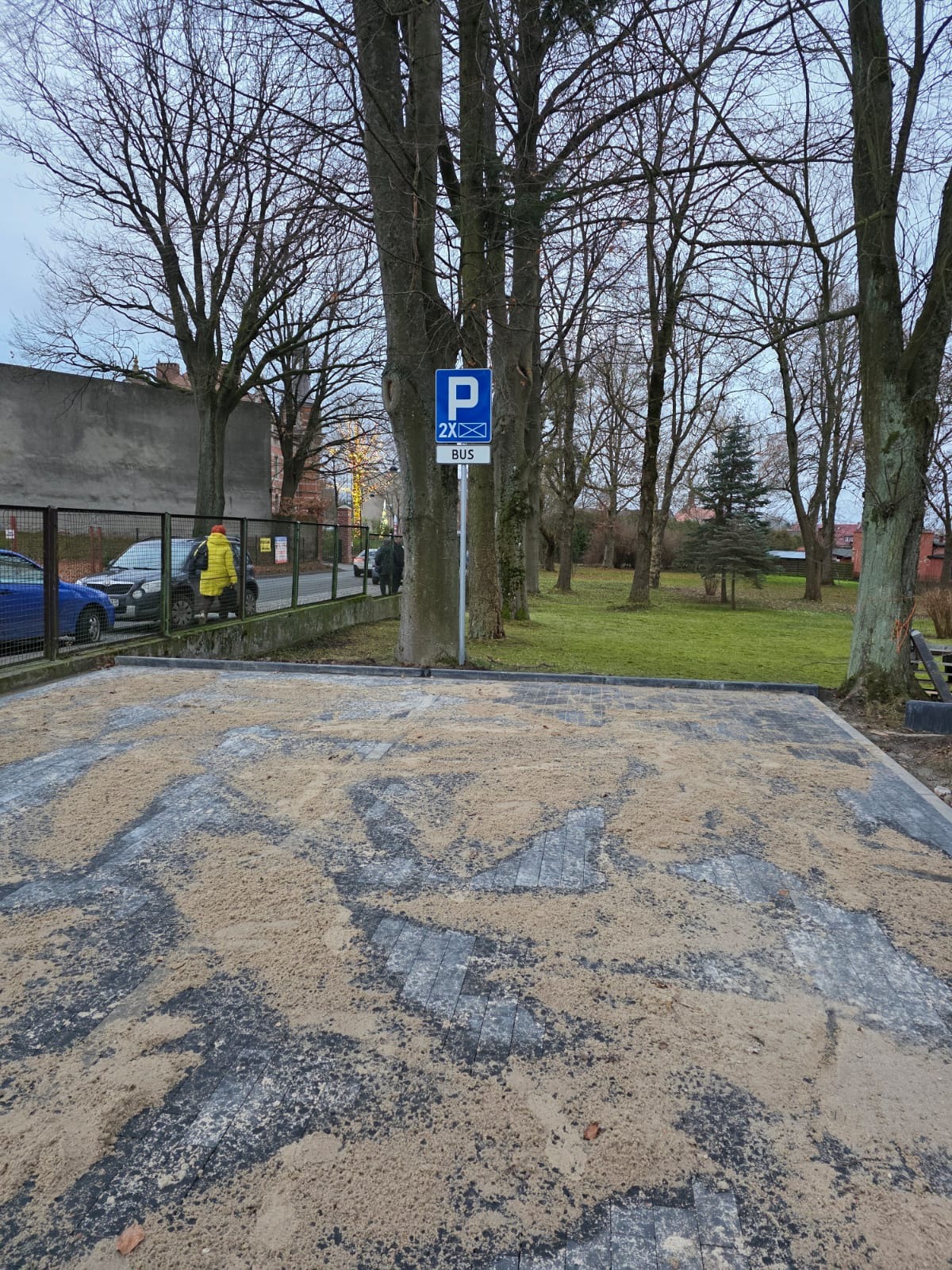 Częściowo ułożona kostka brukowa na parkingu dla autobusów, posypana piaskiem, z widocznym znakiem drogowym i drzewami w tle, w pochmurny dzień.
