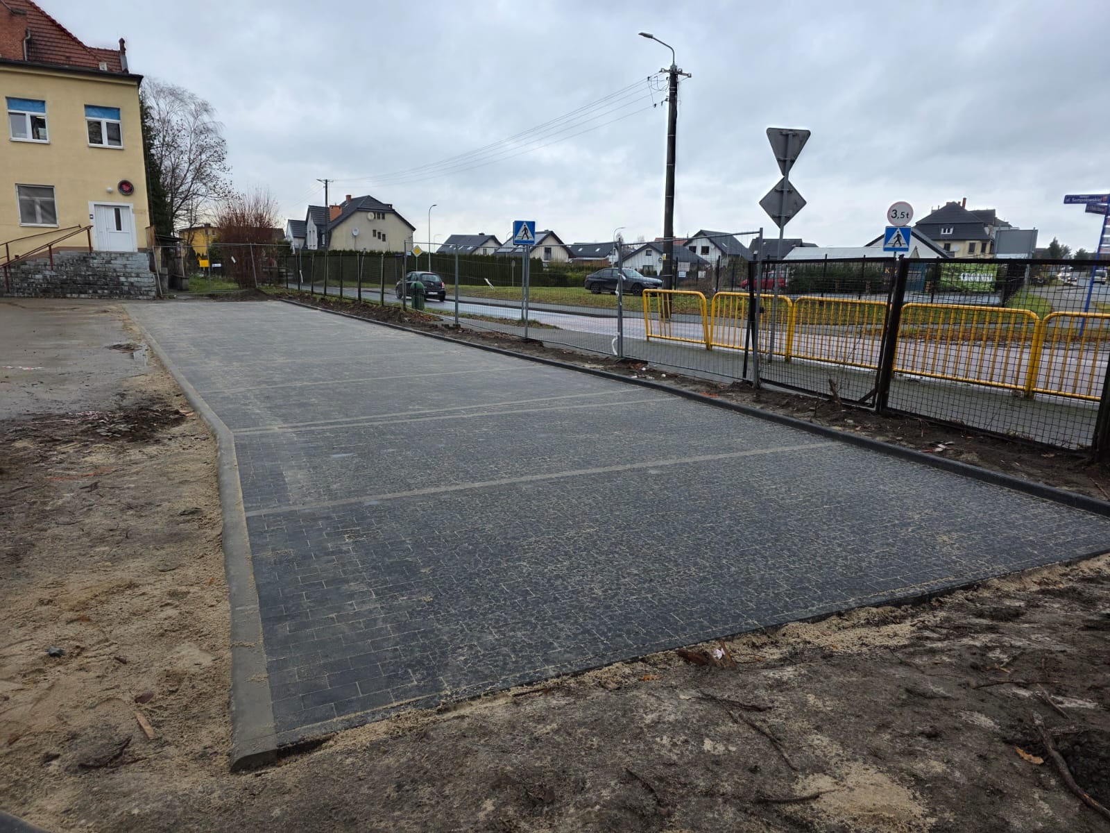 Ukończony parking z kostki brukowej w szarym odcieniu, z widocznymi liniami wyznaczającymi miejsca postojowe. Otoczenie w trakcie prac ziemnych, z widocznym budynkiem w tle.
