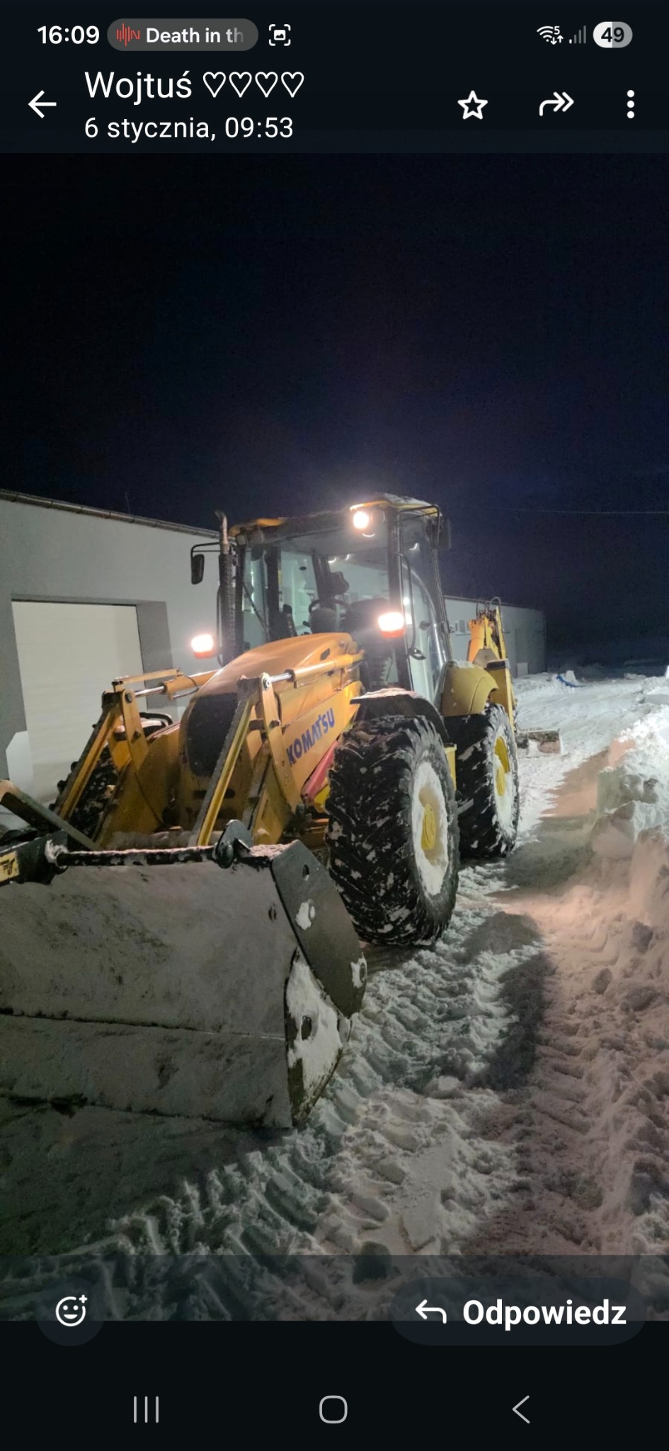 Żółta koparko-ładowarka Komatsu odśnieża teren w nocy, widoczne ślady opon w śniegu i oświetlony budynek w tle.