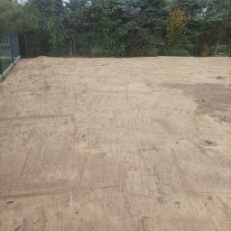 Marcin Czarnecki - Przygotowany teren pod budowę z widocznymi śladami maszyn, otoczony zielenią i ogrodzeniem. W tle dom z szarym dachem i okrągłym oknem.