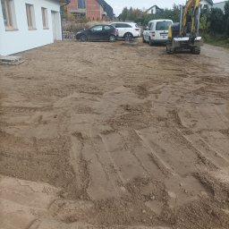 Marcin Czarnecki - Żółta mini koparka na piaszczystym terenie budowy, obok zaparkowane samochody. W tle dom w stanie surowym i budynek z białymi ścianami.