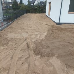 Marcin Czarnecki - Przygotowany teren pod budowę domu jednorodzinnego. Widoczne ślady opon na piasku, ogrodzenie i fragment budynku w tle. Teren wyrównany, gotowy do dalszych prac.