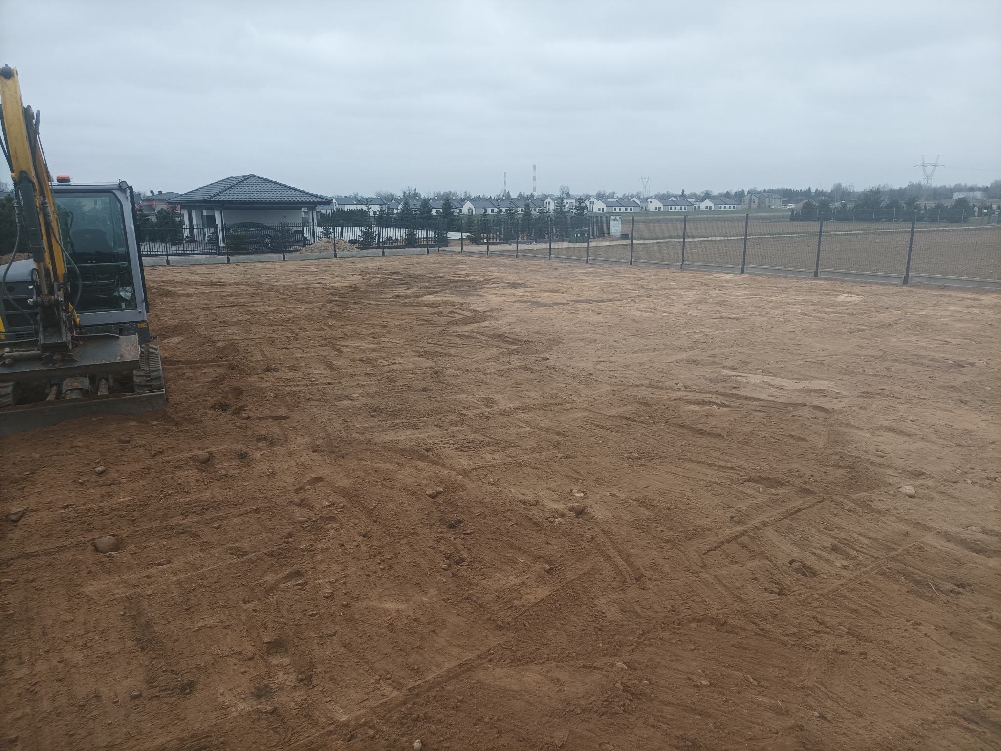 Żółty minikoparka na wyrównanym, brązowym terenie budowlanym, w tle ogrodzenie z siatki, domy jednorodzinne i pochmurne niebo.