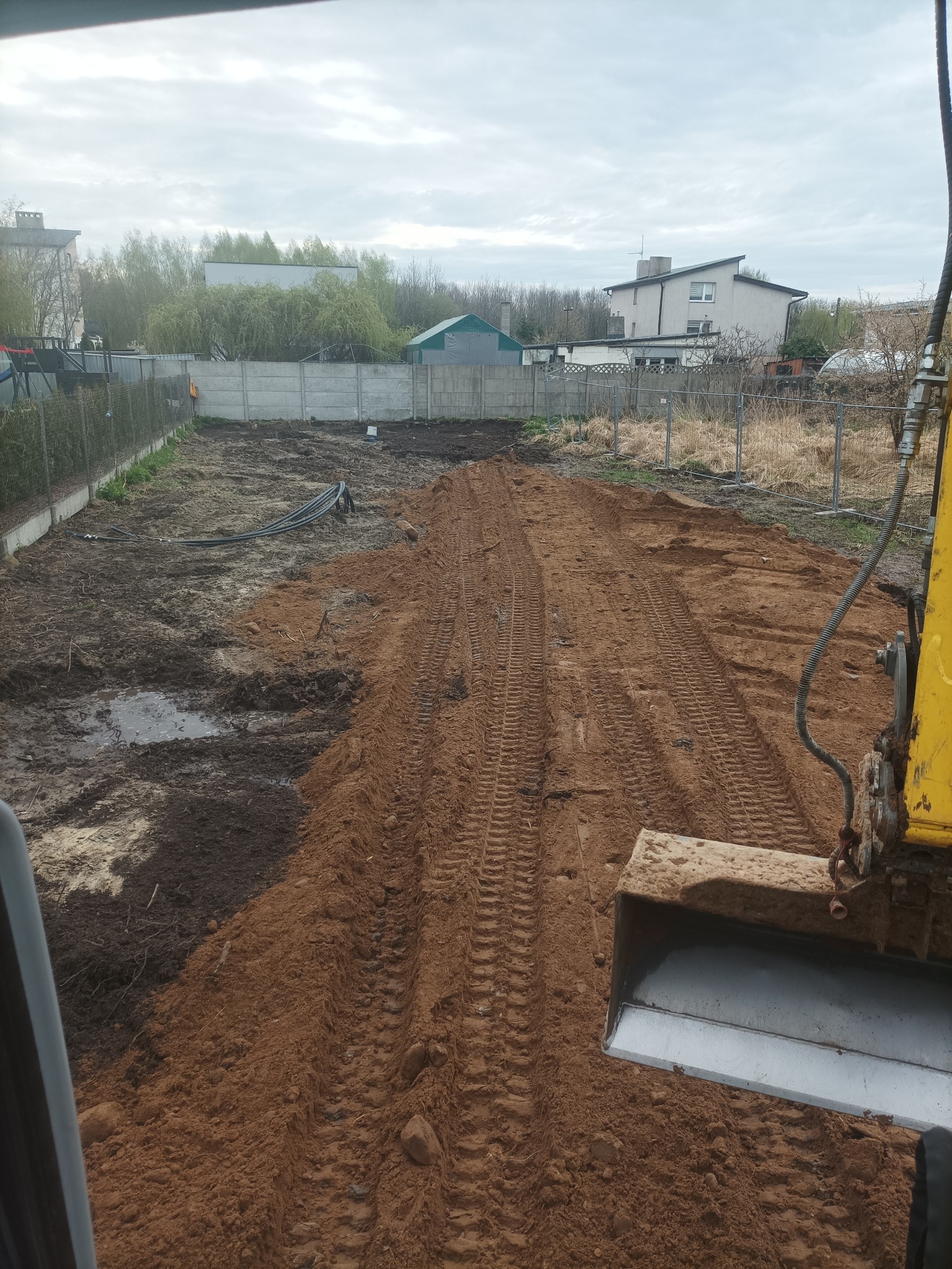 Niwelacja terenu z użyciem koparki, widoczne ślady opon na wyrównanej, czerwonej ziemi, w tle ogrodzenie z siatki i betonowych płyt oraz budynki.