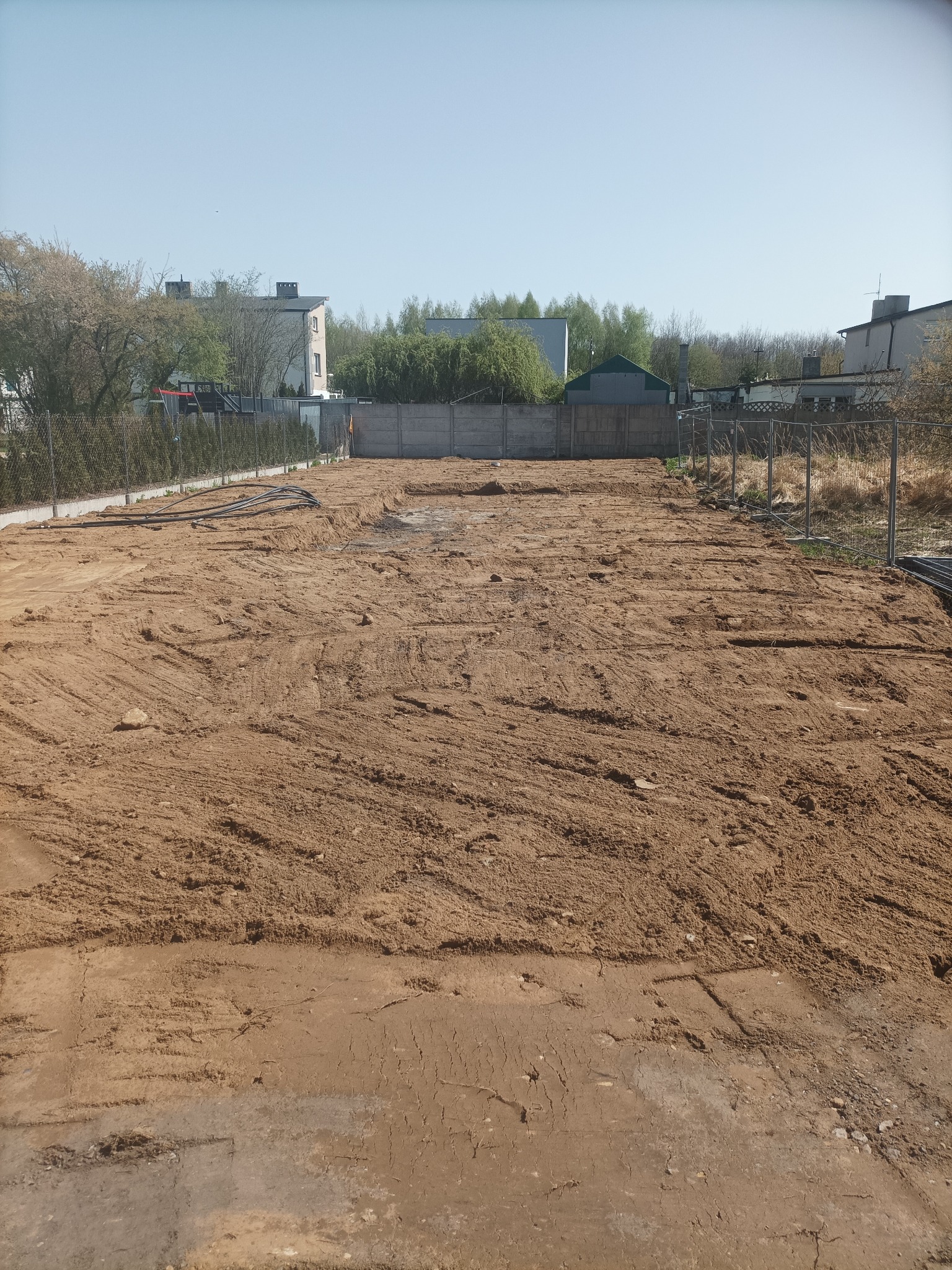 Przygotowany teren pod budowę, z widocznymi śladami maszyn i rurami na powierzchni, ogrodzony siatką i betonowym płotem, w tle zabudowa mieszkalna.