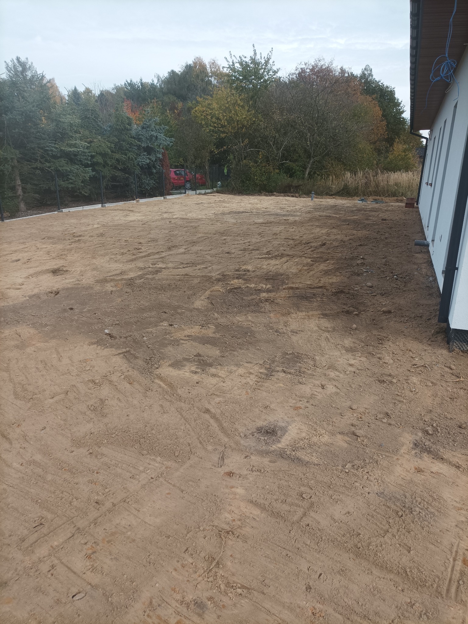 Przygotowany teren pod budowę domu jednorodzinnego, widoczne ślady po pracach ziemnych, w tle drzewa i częściowo widoczny czerwony samochód.