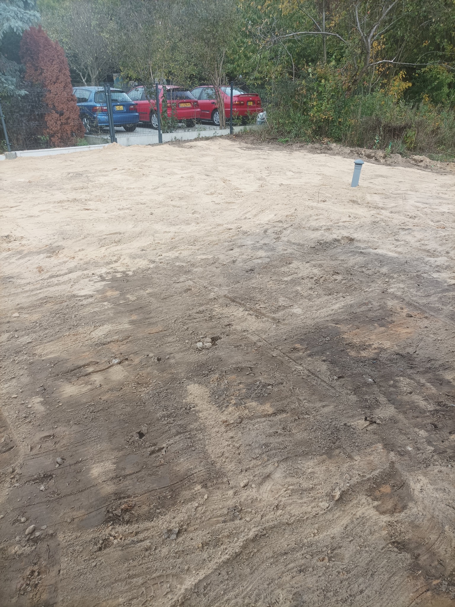 Wyrównany teren pod budowę z widocznymi śladami maszyn, szary przewód wystaje z ziemi. W tle ogrodzenie, drzewa i zaparkowane samochody.