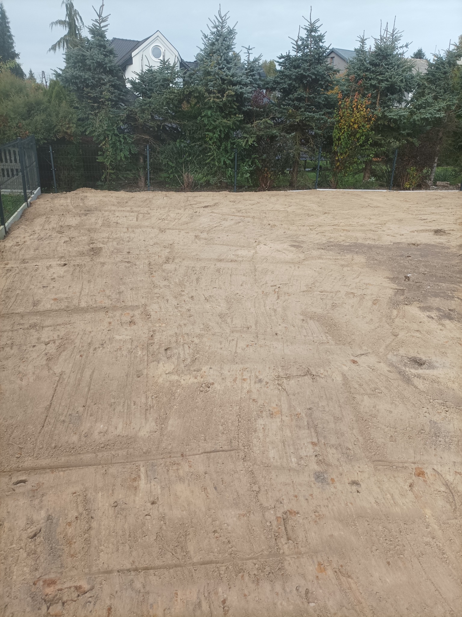 Przygotowany teren pod budowę z widocznymi śladami maszyn, otoczony zielenią i ogrodzeniem. W tle dom z szarym dachem i okrągłym oknem.