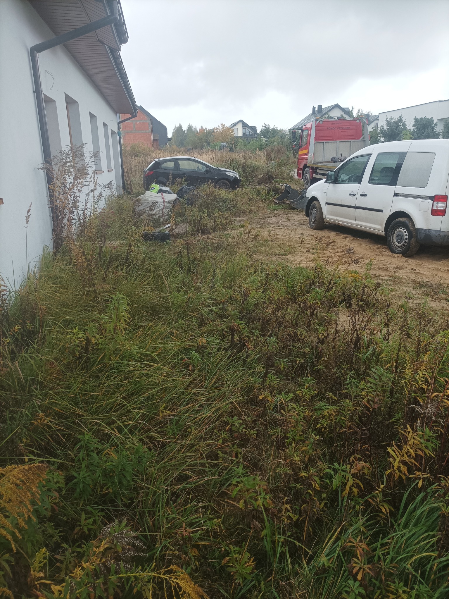 Teren przed budynkiem zarośnięty trawą, widoczny samochód pomocy drogowej i biały van. Przygotowanie terenu pod budowę, wstępny etap prac ziemnych.
