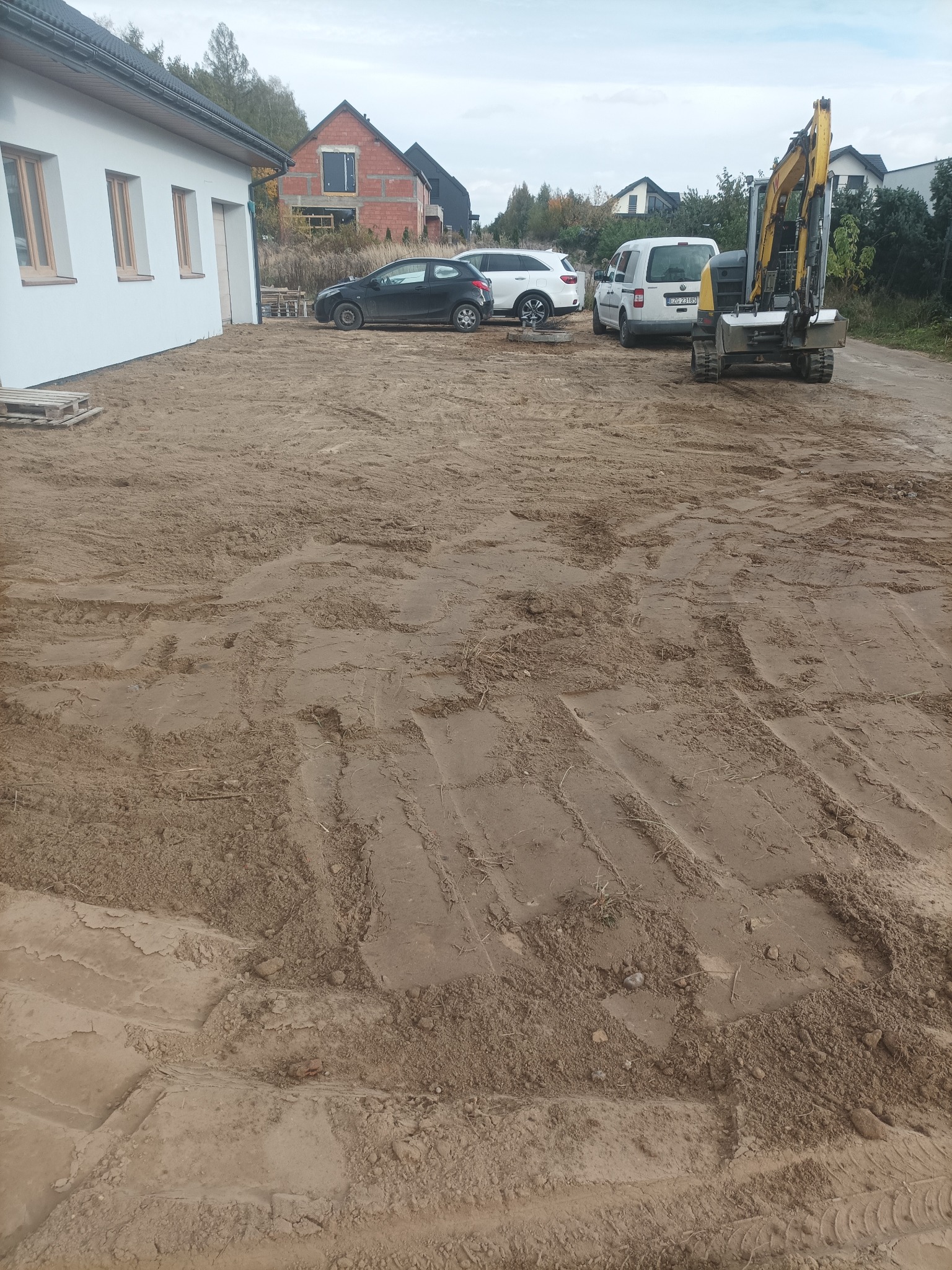 Żółta mini koparka na piaszczystym terenie budowy, obok zaparkowane samochody. W tle dom w stanie surowym i budynek z białymi ścianami.