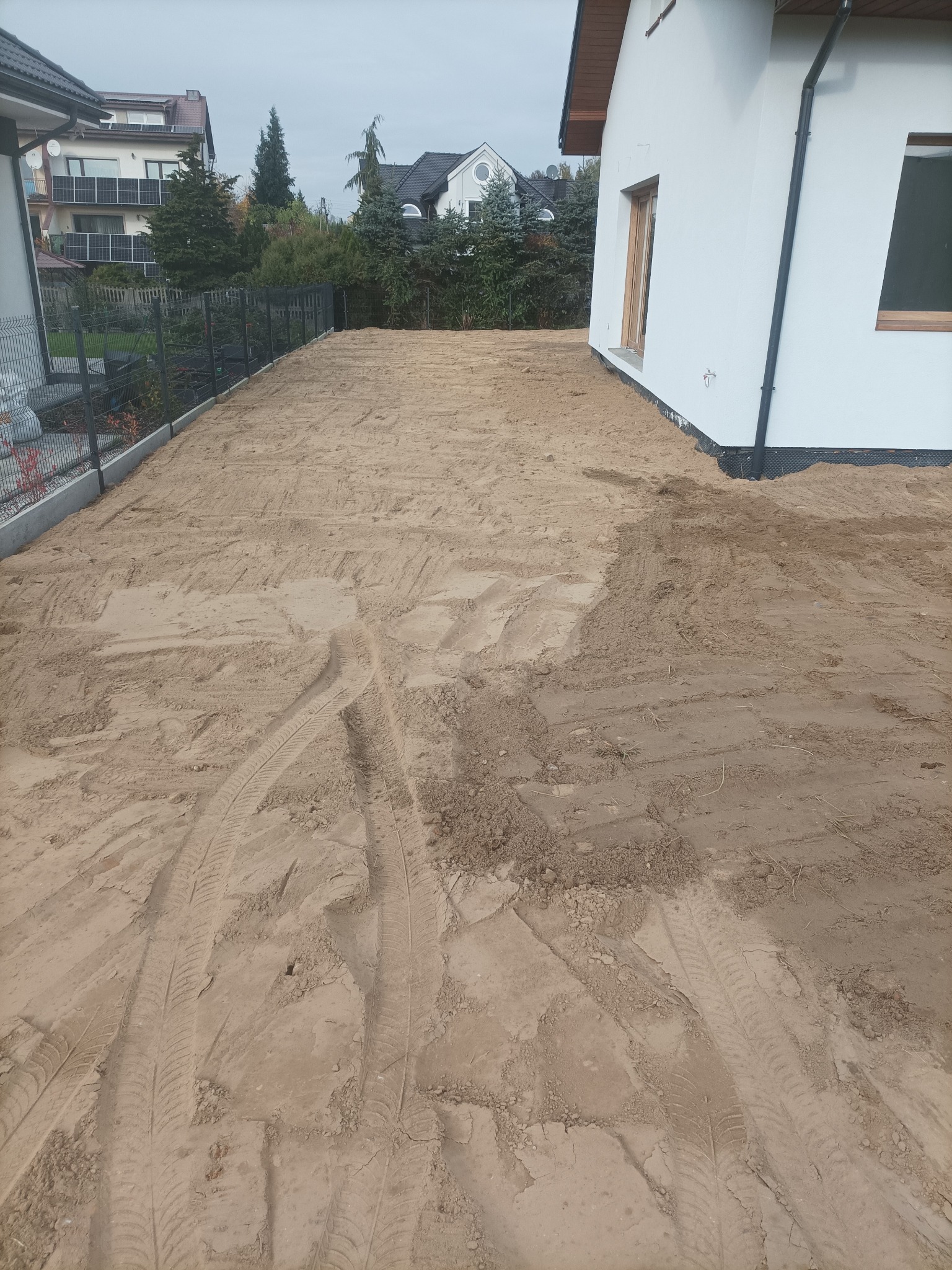 Przygotowany teren pod budowę domu jednorodzinnego. Widoczne ślady opon na piasku, ogrodzenie i fragment budynku w tle. Teren wyrównany, gotowy do dalszych prac.