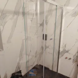 Montaż kabiny prysznicowej z przesuwnymi drzwiami, widoczne marmurowe płytki na ścianach i ciemna podłoga, w tle torba narzędziowa i niebieskie opakowania.