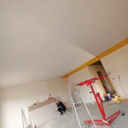 Damian Jurasik - Montaż płyty karton gips na suficie w trakcie prac wykończeniowych, widoczny podnośnik do płyt, drabina i pracownik w trakcie szpachlowania łączeń.