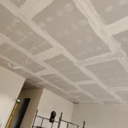 Sufit z płyt kartonowo-gipsowych w trakcie montażu, widoczne łączenia szpachlowane, przewody wystające z sufitu, rusztowanie i lampa budowlana w pomieszczeniu.