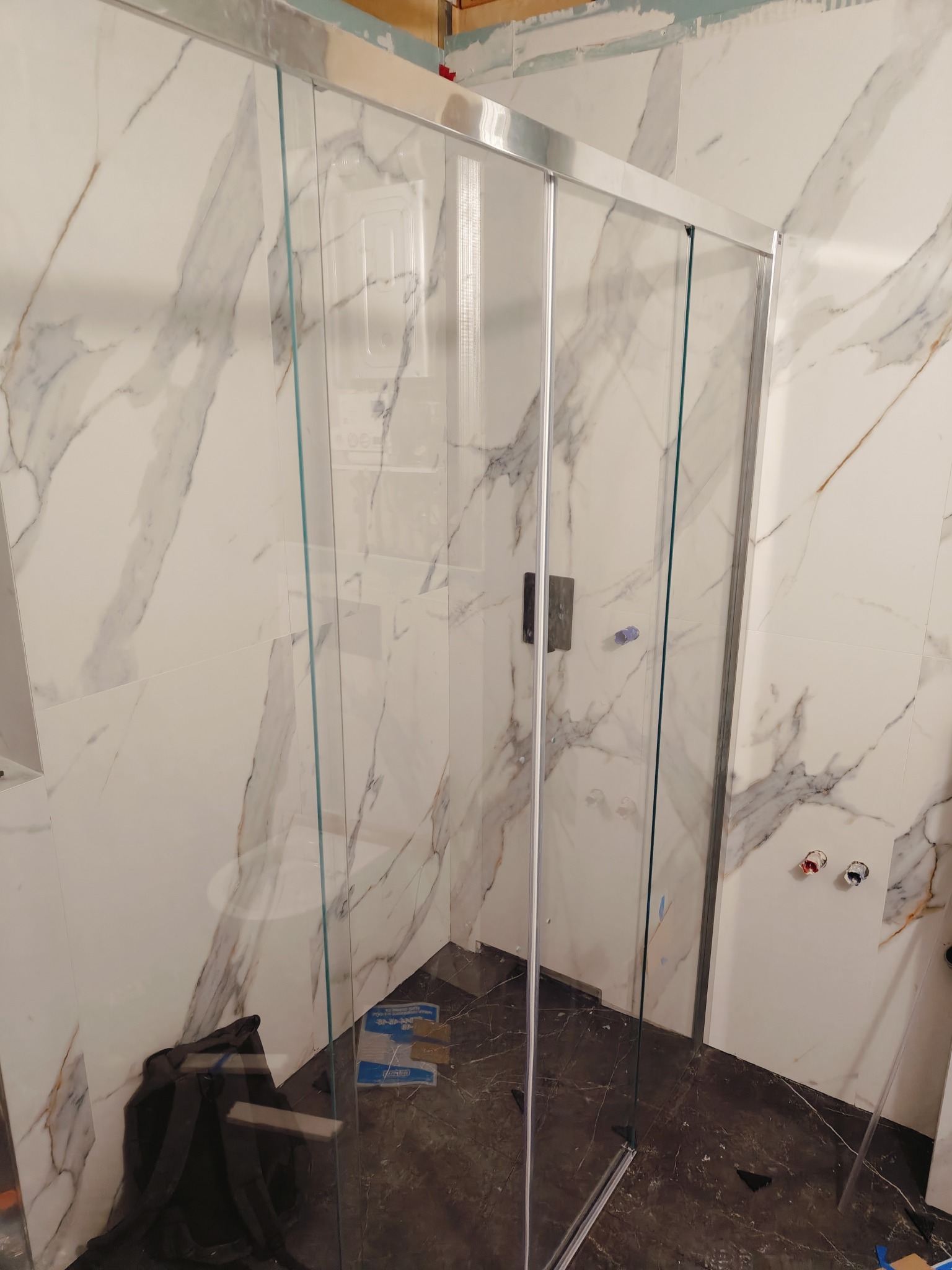 Montaż kabiny prysznicowej z przesuwnymi drzwiami w łazience wyłożonej białymi płytkami imitującymi marmur, widoczne narzędzia i materiały budowlane na podłodze.