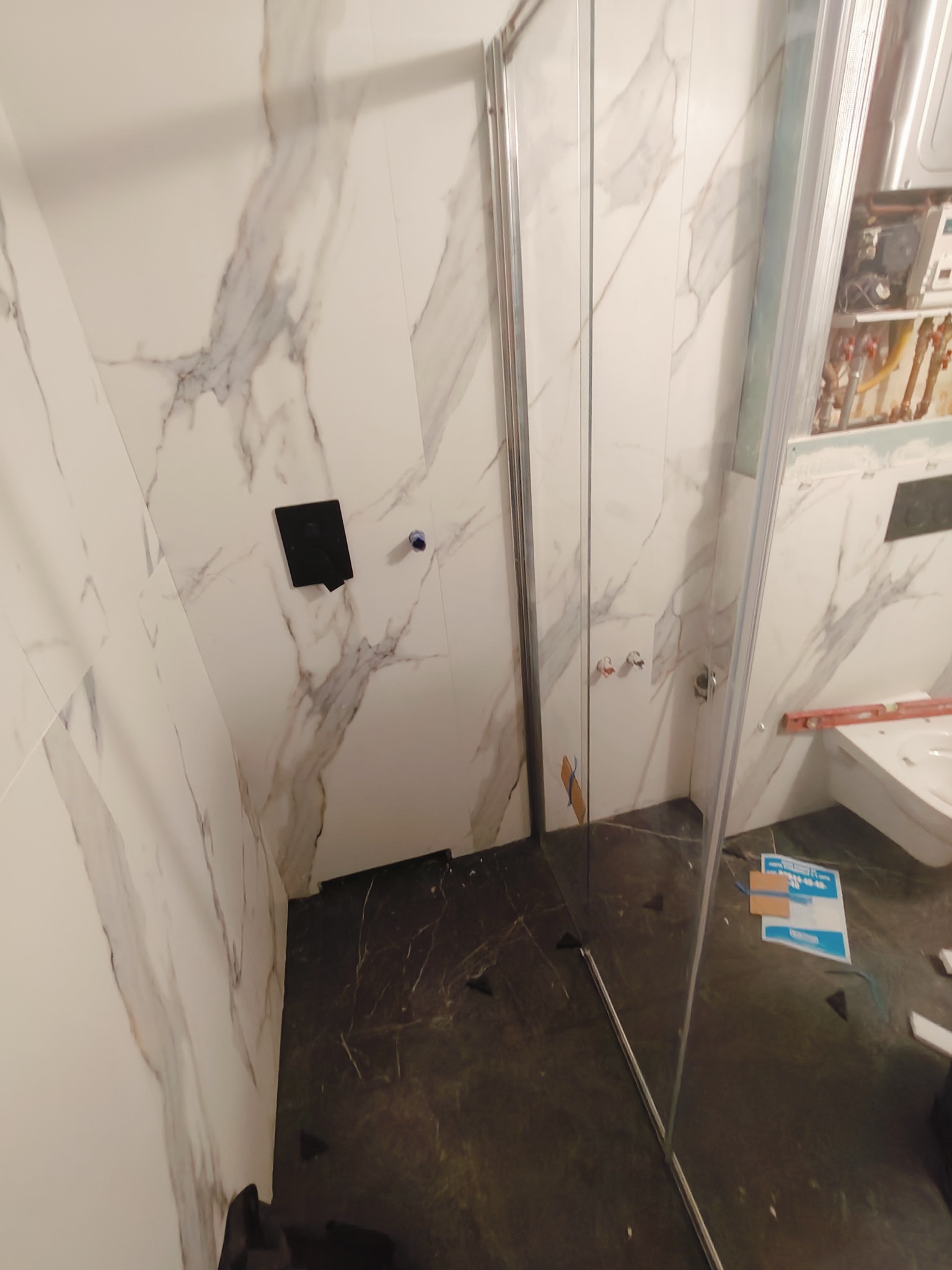 Montaż kabiny prysznicowej z białymi płytkami imitującymi marmur i czarną podłogą w trakcie prac wykończeniowych, widoczny fragment instalacji wodnej i poziomica.