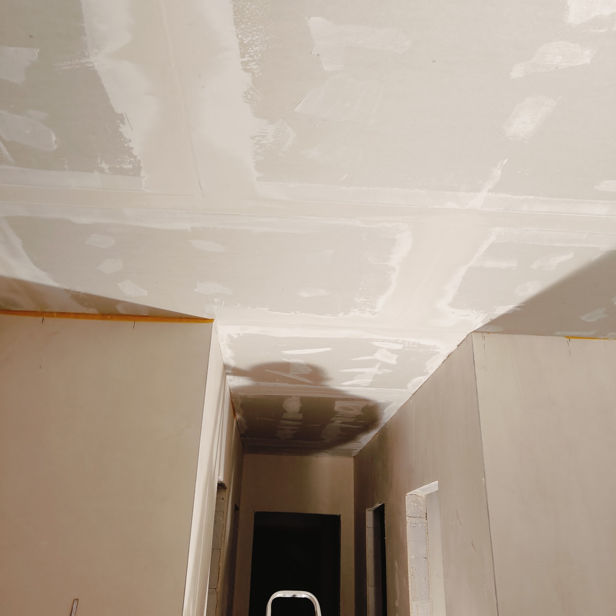 Sufit i ściany po szpachlowaniu, widoczne łączenia płyt karton-gips, przygotowane do malowania, żółta taśma malarska przy suficie, w tle widoczny fragment drabiny.