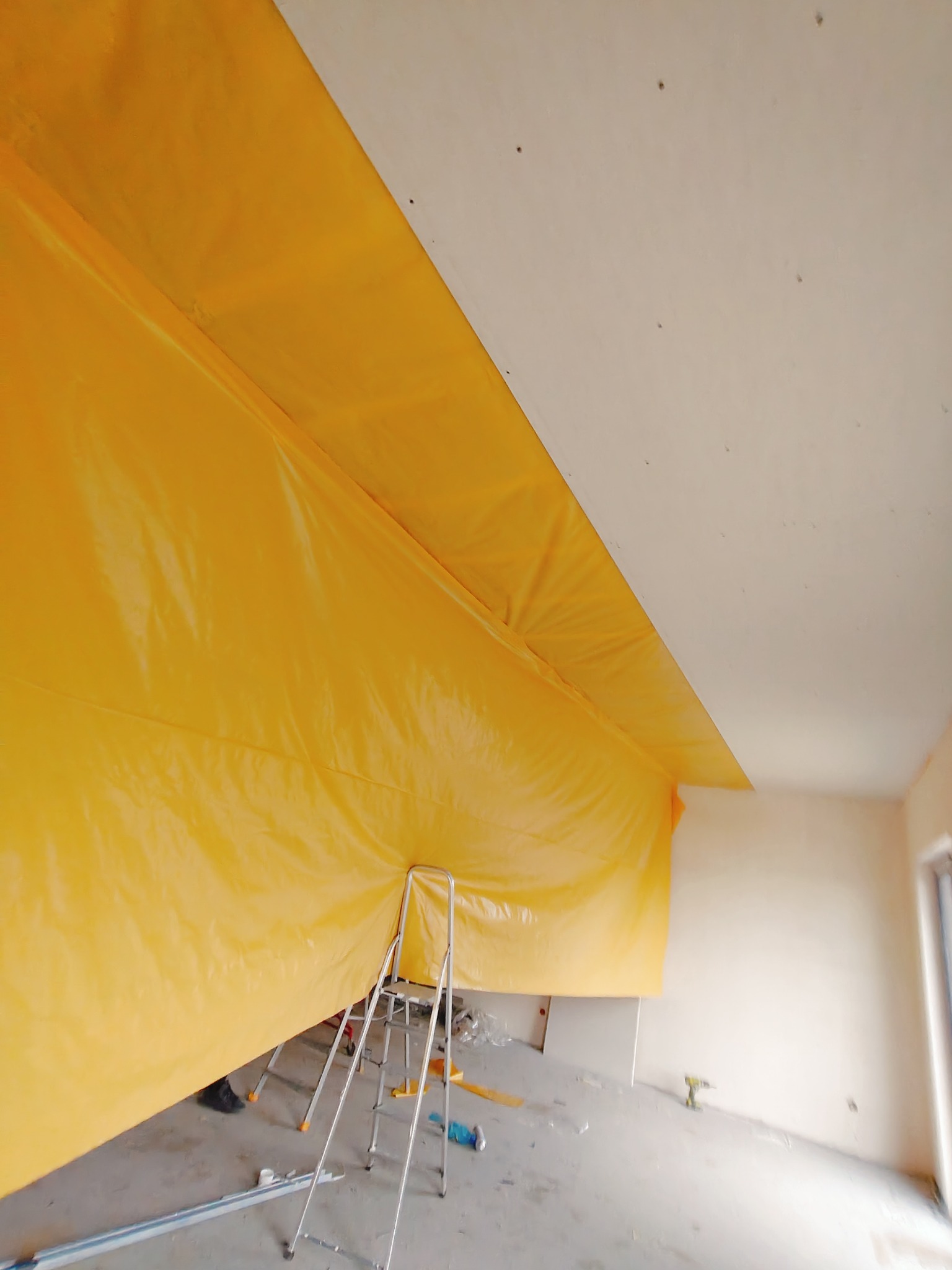 Przygotowanie do montażu sufitu podwieszanego: folia ochronna na ścianie, drabina aluminiowa i wkrętarka akumulatorowa na podłodze.