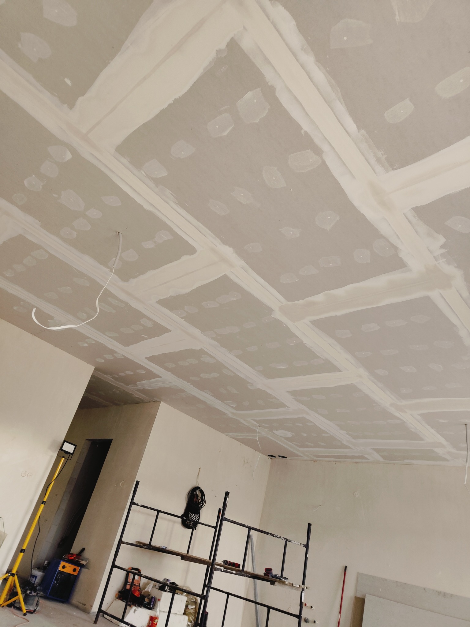 Sufit z płyt kartonowo-gipsowych w trakcie montażu, widoczne łączenia szpachlowane, przewody wystające z sufitu, rusztowanie i lampa budowlana w pomieszczeniu.