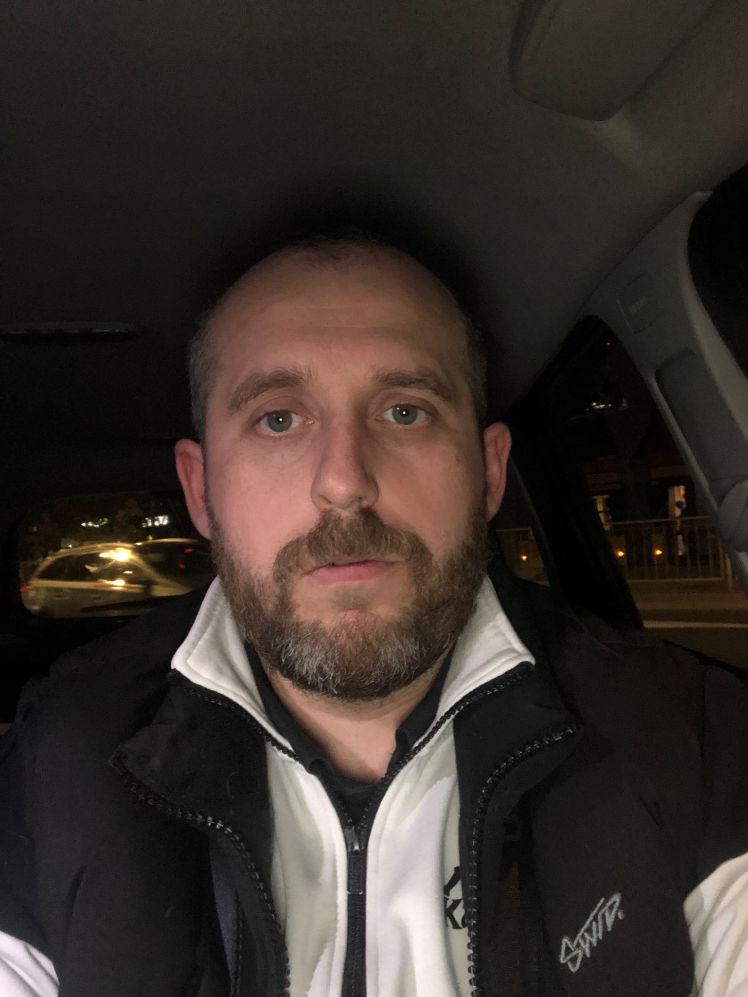 Mężczyzna z brodą w samochodzie, widok z perspektywy pasażera, w tle światła uliczne i fragment innego pojazdu.