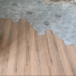 Kyrylo Homeniuk - Układanie jasnych paneli podłogowych obok szarych, heksagonalnych płytek podłogowych w trakcie remontu.