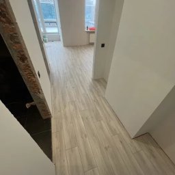 Kyrylo Homeniuk - Jasne panele podłogowe ułożone wzdłuż korytarza, widok na okno i fragment ściany z surową cegłą po lewej stronie.