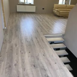 Kyrylo Homeniuk - Jasnoszare panele podłogowe imitujące drewno, ułożone w nowym salonie z widokiem na schody z podświetleniem LED.